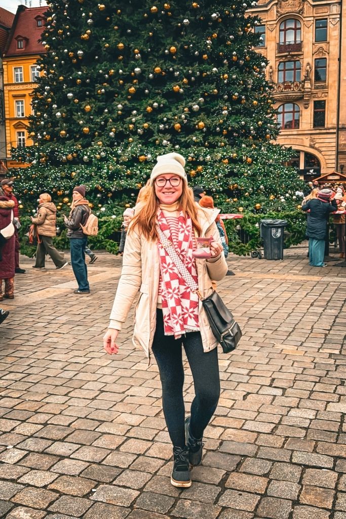 Kate wearing a cream winter coat, white knit beanie, red-and-white patterned scarf, black leggings, and black boots in front of a giant Christmas tree in Wroclaw.