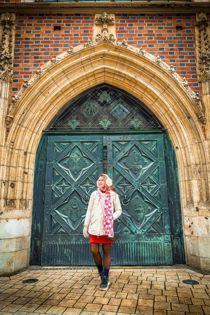 Kate wearing a cream winter coat, red-and-white patterned scarf, red skirt, black tights, and black winter boots in front of an ornate historic doorway in Wroclaw.